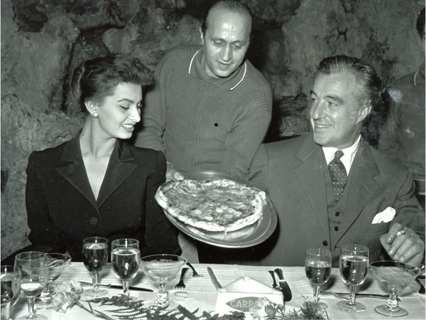 foto Ezio Vitale, Sophia Loren e Vittorio De Sica accolgono, sorridenti, una gustosa pizza napoletana, 1960 circa, bianco e nero. Al ristorante Vesuvio di Napoli