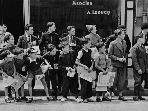 Robert Capa, France, Brittany, Pleyben, July 1939. A crowd gathered in front of Mr. Pierre Cloarec's bicycle shop. The owner of the shop is racing in the Tour de France