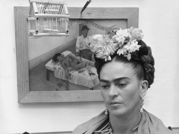 FRIDA. VIVA LA VIDA, La pittrice messicana Frida Kahlo (1907 - 1954) siede con le braccia conserte, guardando in basso, di fronte a uno dei suoi dipinti e una gabbia per uccelli in legno. Indossa fiori tra i capelli e una collana di legno | Foto: Hulton Archive - Getty Images | Courtesy of Ballandi Arts e Nexo Digital 2019