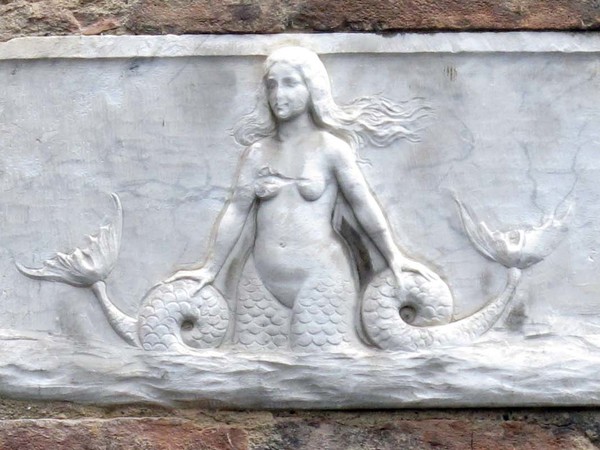 Targa con Sirena bicaudata, Palazzetto in Via dei Rossi, Siena | foto: Sailko (Opera propria) via Wikimedia Creative Commons | La sirena a due code di epoca medievale scolpita in moltissimi luoghi di culto e non solo, risale ad una simbologia millenaria legata all'antichissimo Culto della Dea Madre diffuso nell’Europa Neolitica oltre che ai più recenti culti di alcune divinità egizie come Api e Bastet.